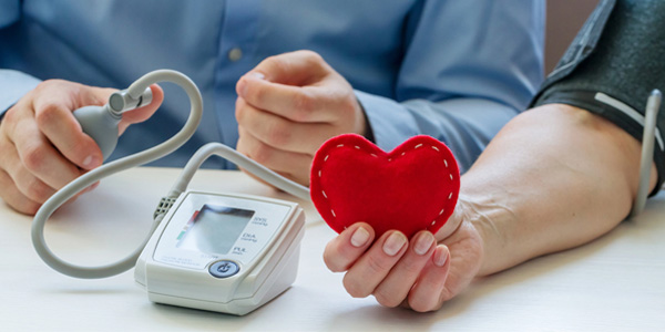 Arm mit Blutdruckmanschette neben einem Blutdruckmessgerät, in der Hand ein rotes Stoffherz.